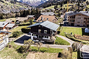 Aerial view of a dark-wood chalet amidst a sunny mountain village with green hills and snow-capped peaks in the background.