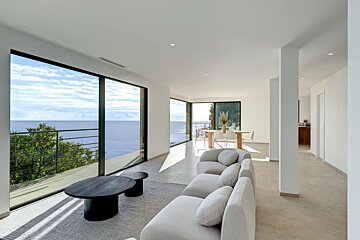 Bright, modern living room with panoramic ocean views through large windows, featuring light-colored sofas and a minimalist aesthetic.
