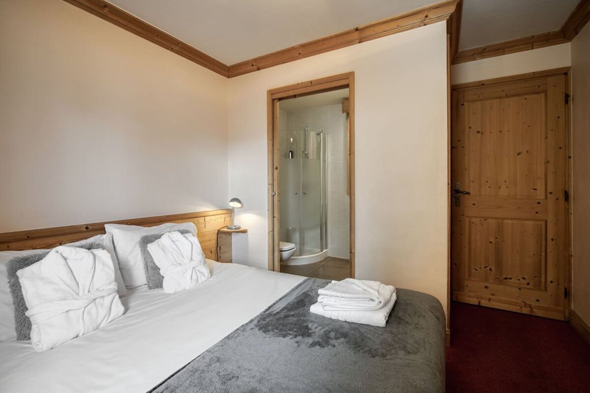 Cozy bedroom with white bed, grey throw, bathrobes, and wooden accents. An open doorway leads to a bathroom with a shower, plus a wooden door.