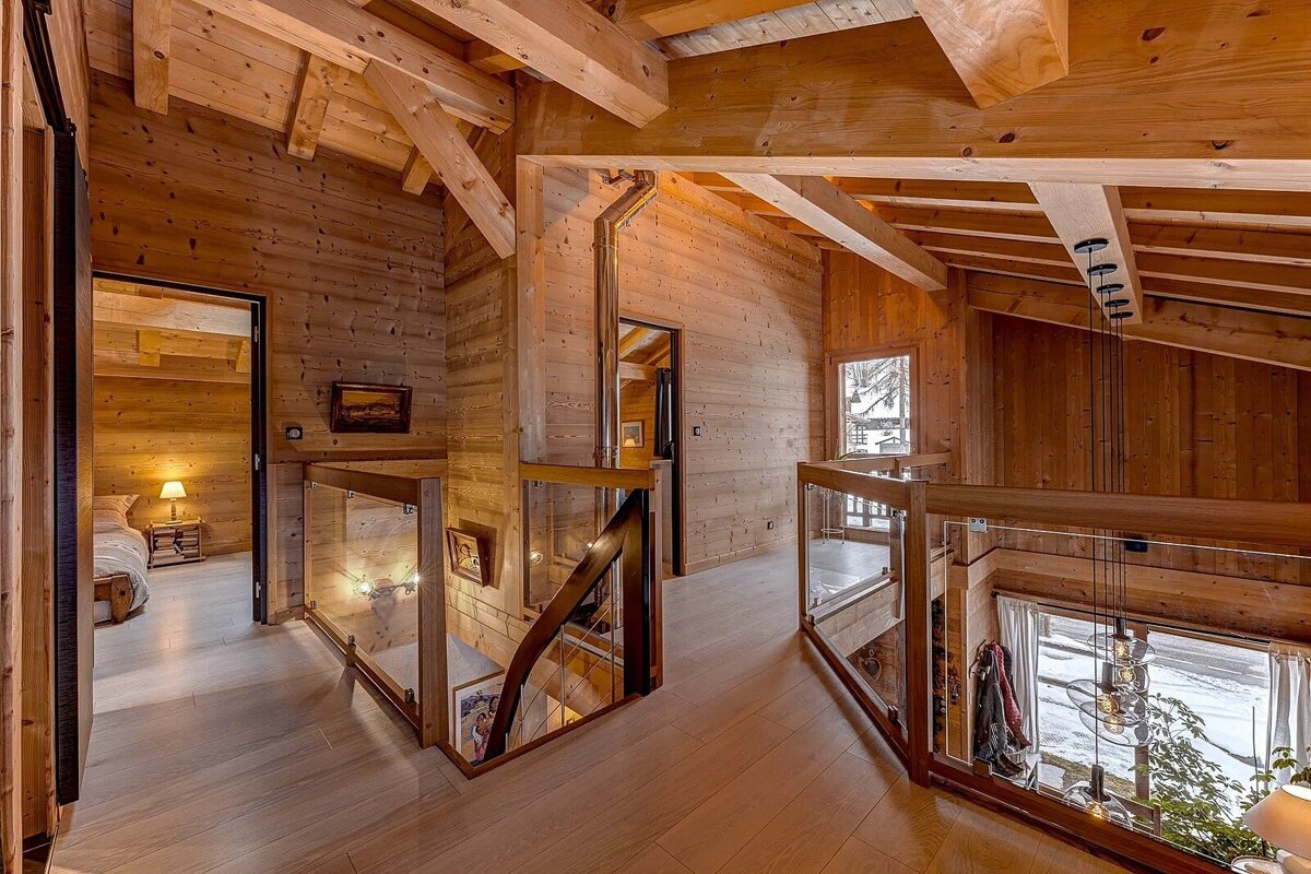 A warm, multi-level wooden chalet interior with exposed beams, glass railings, hallways, and a bedroom, offering a snowy outdoor view.