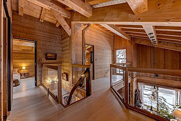 A warm, multi-level wooden chalet interior with exposed beams, glass railings, hallways, and a bedroom, offering a snowy outdoor view.