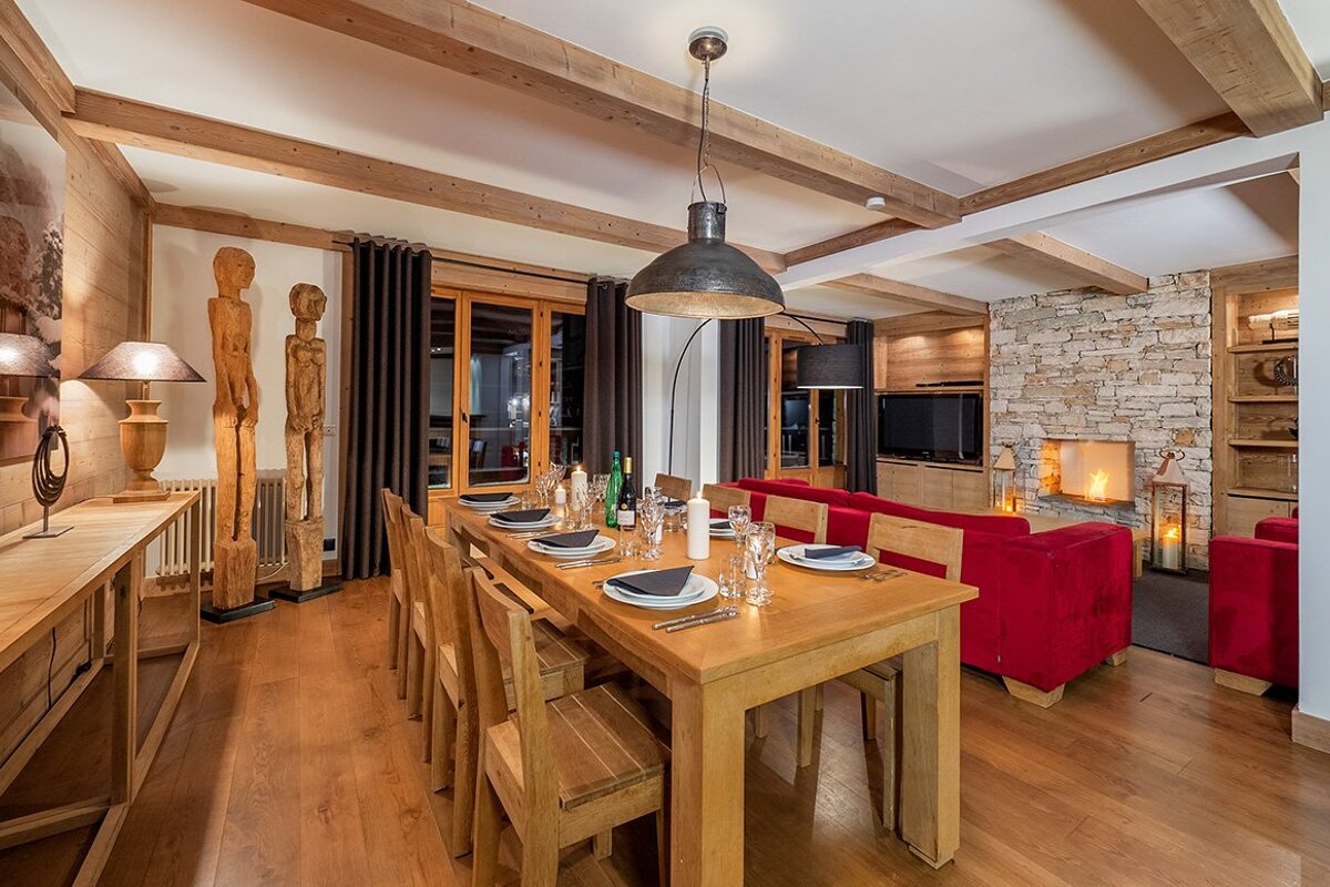 Cozy chalet-style open concept living and dining room with a long wooden table, red sofas, stone fireplace, and wooden accents.