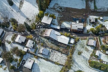 Aerial view of a rural area lightly dusted with snow. White-roofed houses, roads, and green trees dot the landscape with patches of snowy and muddy ground.