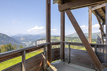Panoramic mountain view from a wooden structure. Green valleys, distant peaks, and a clear blue sky on a sunny day.