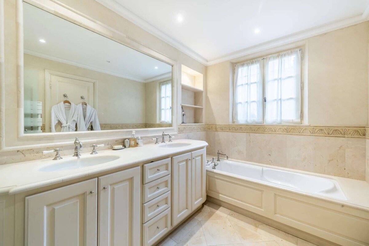 A luxurious, cream-toned bathroom features a double vanity with a large mirror, a bathtub, and a window with sheer curtains. Two robes hang in the mirror's reflection.