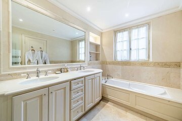 A luxurious, cream-toned bathroom features a double vanity with a large mirror, a bathtub, and a window with sheer curtains. Two robes hang in the mirror's reflection.