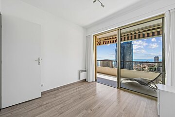 An empty room with white walls, wood floors, a door, and a sliding glass door opening to a balcony with city views.
