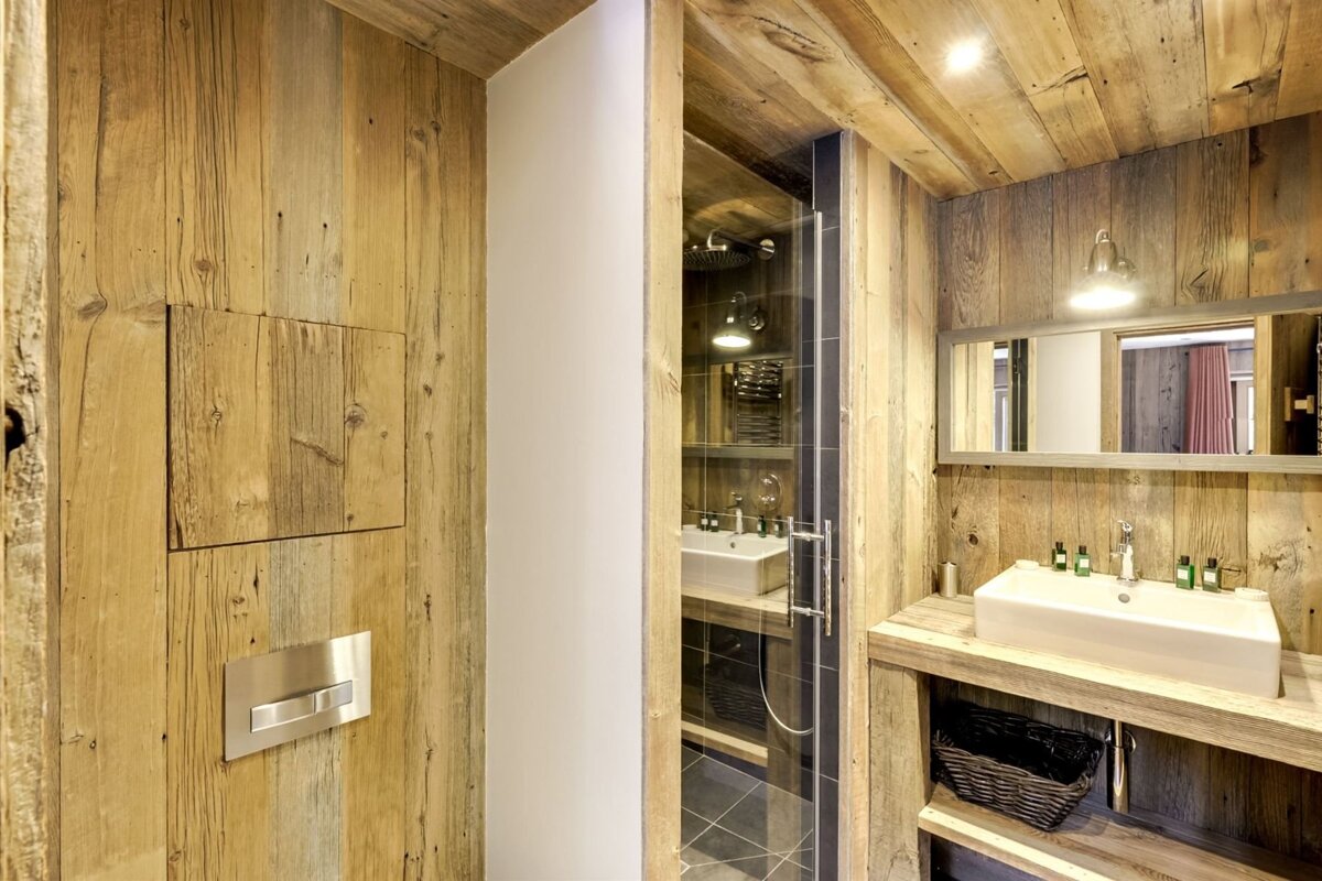 Rustic bathroom with wood paneling. Features a toilet and a modern sink/shower area behind a glass door.