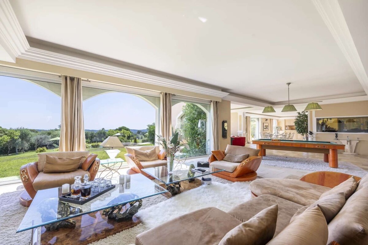 A spacious, luxurious living room with panoramic garden views, plush seating, a glass coffee table, and a pool table in the background.