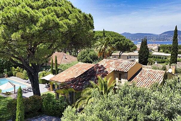 Aerial view of a Mediterranean town with red-tiled roofs, lush trees, a pool, and a blue sea with mountains in the background under a clear sky.