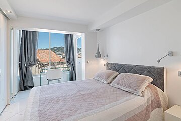 A bright, modern bedroom with a grey headboard, pink quilted bedding, and a large window overlooking scenic tiled rooftops.