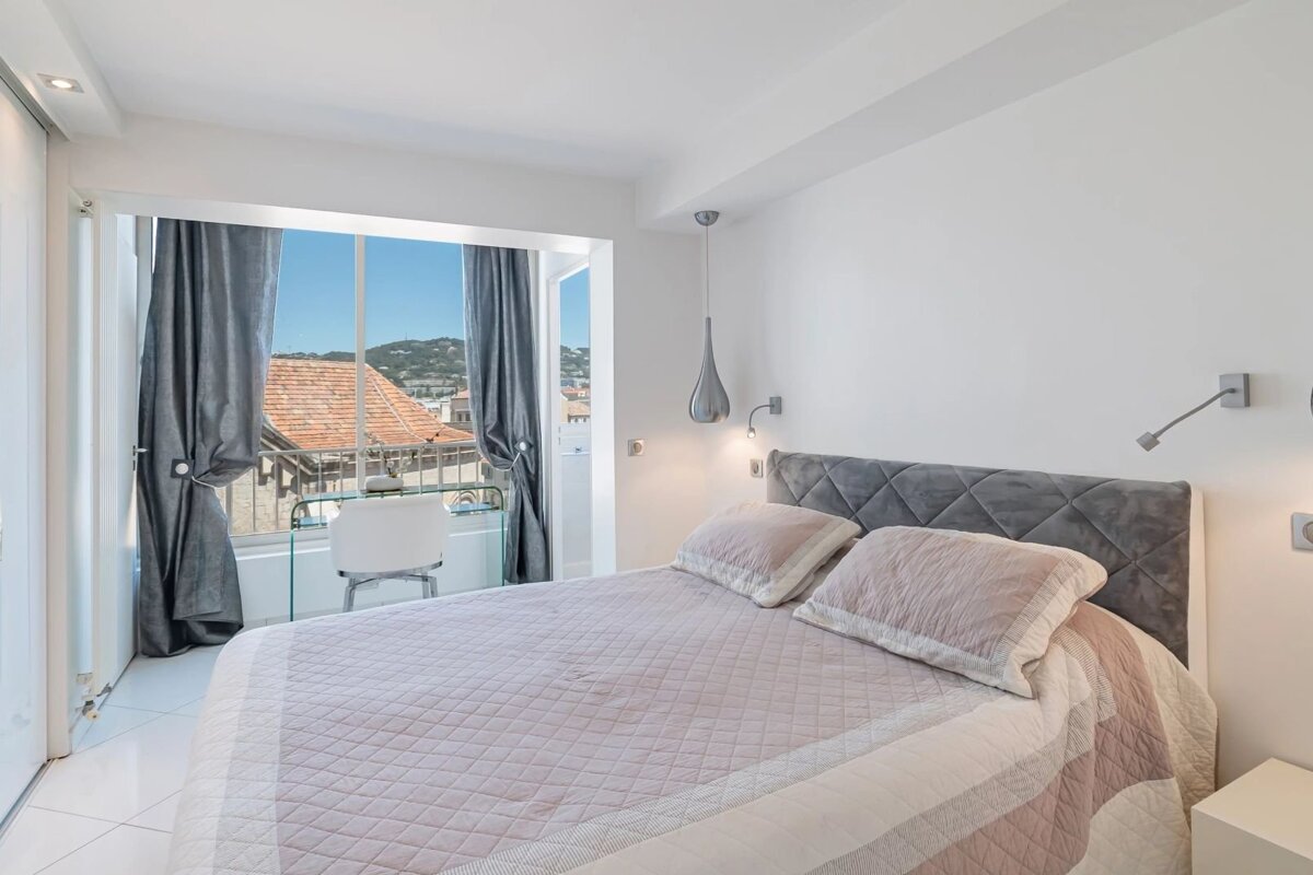 A modern, bright bedroom featuring a quilted pink bed, gray tufted headboard, and a large window with scenic outdoor views.