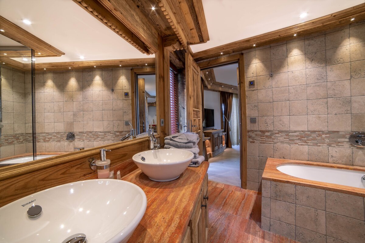 Luxurious, rustic bathroom with warm wood accents, light tiled walls, dual basin vanity, and a built-in tub, well-lit and inviting.