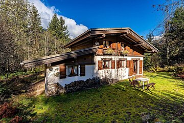 A sunny wooden chalet with brown shutters, stacked firewood, and an outdoor table on a green lawn, surrounded by a dense forest under a blue sky.