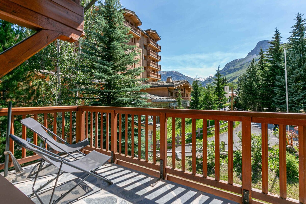 A balcony with lounge chairs offers a mountain resort view featuring wooden chalets, tall pines, and distant peaks under a bright blue sky.