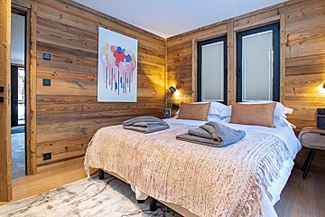 Cozy rustic bedroom with wooden paneled walls, a neatly made bed with grey folded blankets, and colorful art on the wall.