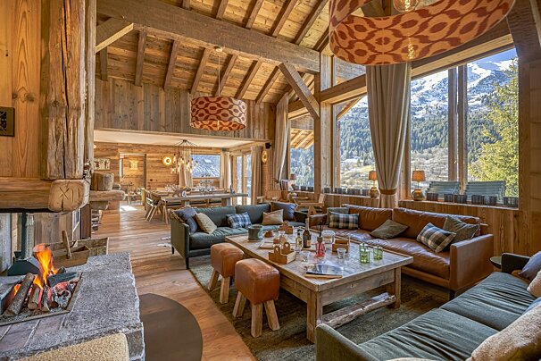A cozy, spacious wooden chalet with a roaring fireplace, comfortable sofas, and panoramic windows framing stunning snowy mountain and forest views.