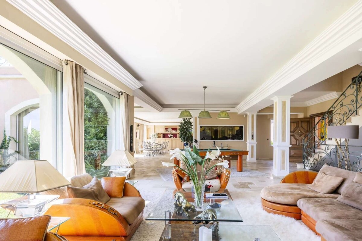 Luxurious, spacious living room with large windows, ornate sofas, a billiard table, dining area, and grand staircase, all under high ceilings.
