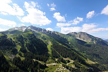 Vibrant summer mountain landscape features towering green peaks, some with snow, dense pine forests, ski slopes, and scattered buildings under a clear blue, cloudy sky.