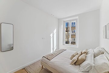 A minimalist white bedroom with a bed, a window showing city buildings, and a mirror. Sunlight streaks across the wall.