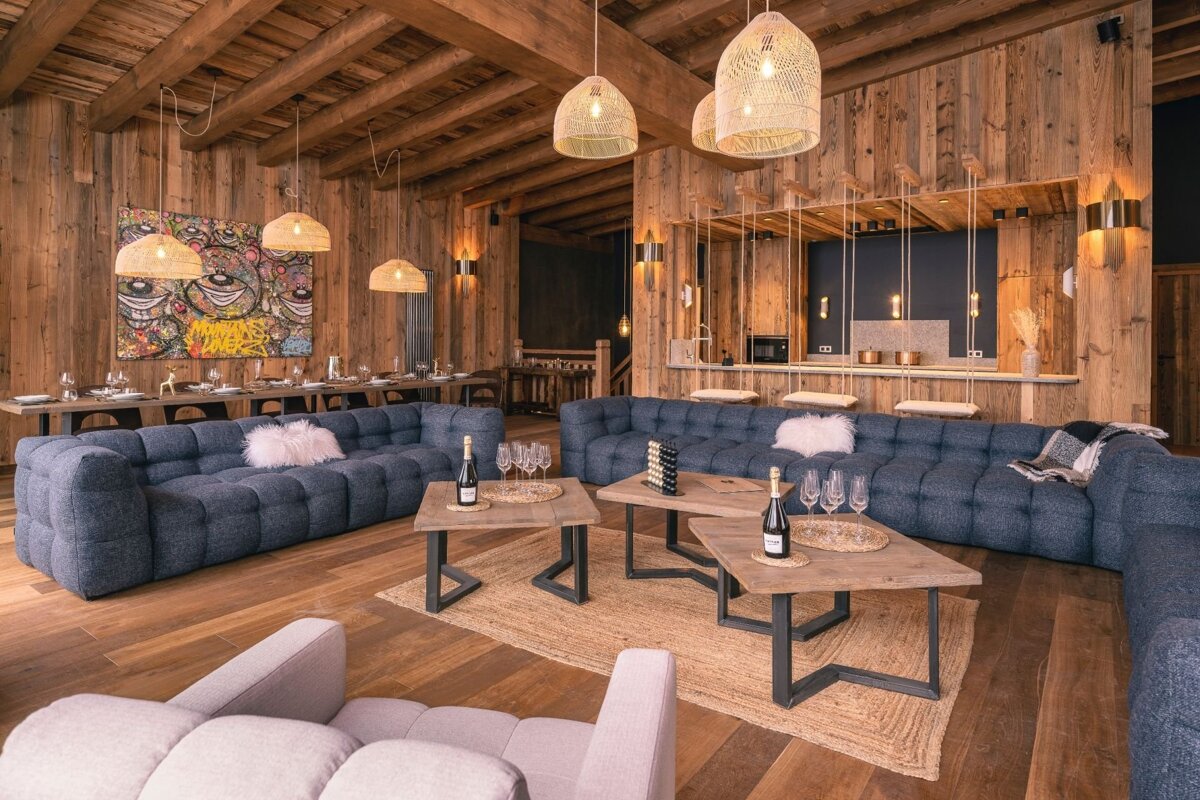 Warm, expansive chalet living area with rich wooden walls, comfy blue sofas, a dining table, and a modern bar.