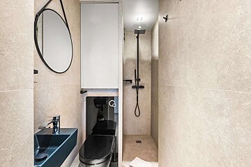 Compact, modern bathroom with light tiles, black toilet, blue sink, black shower fixtures, and a round mirror.