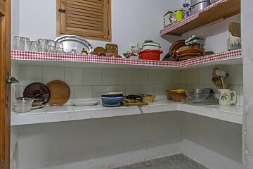 A rustic kitchen pantry with white walls, wooden shutters, and open shelves full of dishes, glasses, and cookware, accented with red checkered liners.