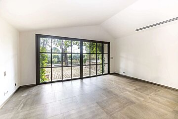 An empty room with white walls, grey tiled floor, and a large black-framed window overlooking a green garden courtyard.