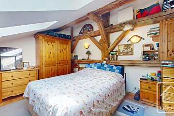Rustic attic bedroom featuring prominent wooden beams, a patterned bed, wooden wardrobe & dresser.