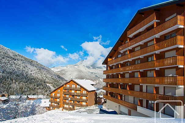A winter scene featuring large wooden chalets nestled in a snow-covered mountain valley, set against a bright blue sky with scattered clouds.