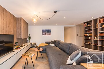 A cozy living room with a large grey sectional sofa, wooden paneling, a TV, and a built-in bookshelf.