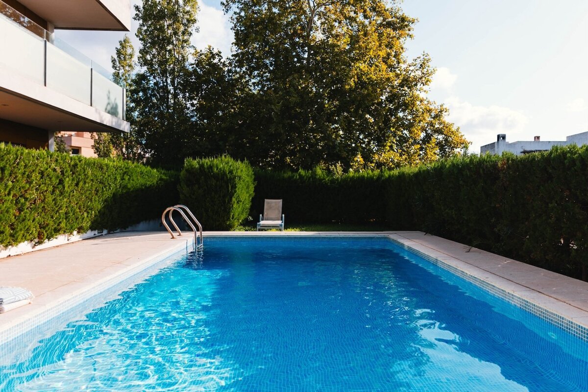A sunny outdoor swimming pool with bright blue water, bordered by green hedges and large trees, next to a modern building with a balcony.