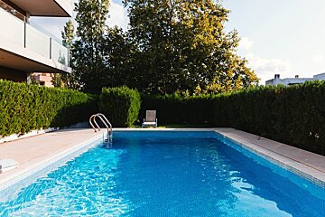 A sunny outdoor swimming pool with bright blue water, bordered by green hedges and large trees, next to a modern building with a balcony.