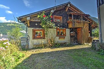 A charming rustic chalet with a wooden balcony and red roses climbing its wall, set against a backdrop of green mountains under a sunny blue sky.