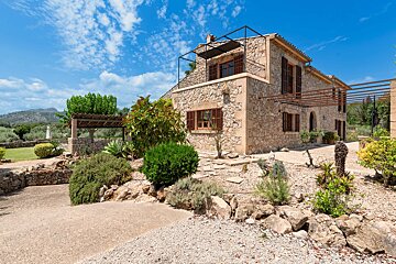 A two-story stone house with brown shutters and a balcony, nestled in a rocky garden with shrubs, under a blue sky, with mountains in the distance.