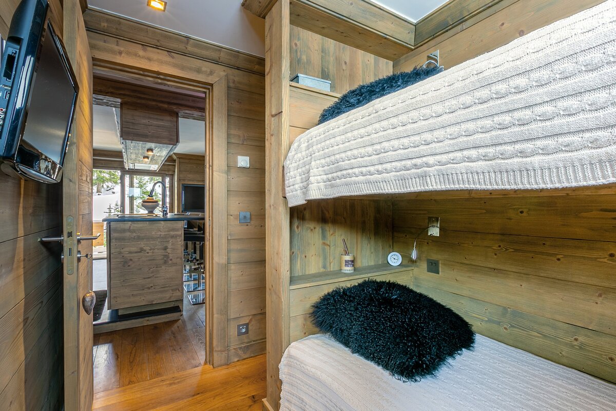 Cozy rustic room with built-in wooden bunk beds, black fluffy pillows, and a glimpse into a modern kitchen/living area through an open doorway.