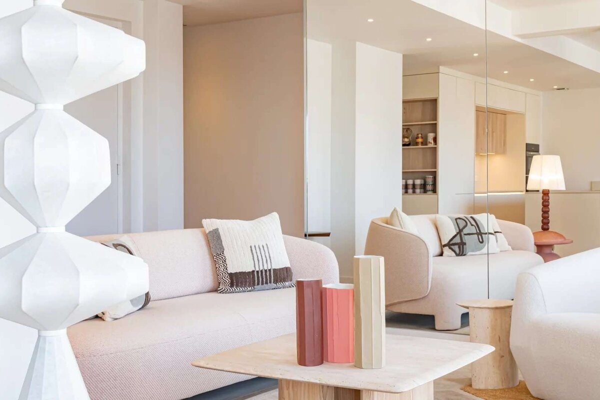 A modern, minimalist living room with pale pink sofas, a unique white lamp, and a coffee table adorned with colorful vases.
