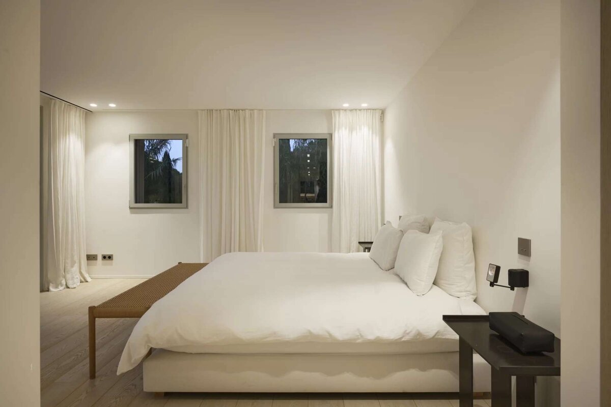 A clean, minimalist bedroom features a large white bed, two windows with sheer curtains, light wooden floors, and a simple wooden bench.