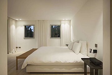A clean, minimalist bedroom features a large white bed, two windows with sheer curtains, light wooden floors, and a simple wooden bench.