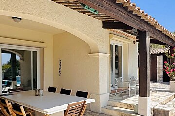 Sunny outdoor patio with dining table, sliding doors, and a pergola. Light-colored building with a tiled roof, and a pool reflection visible.