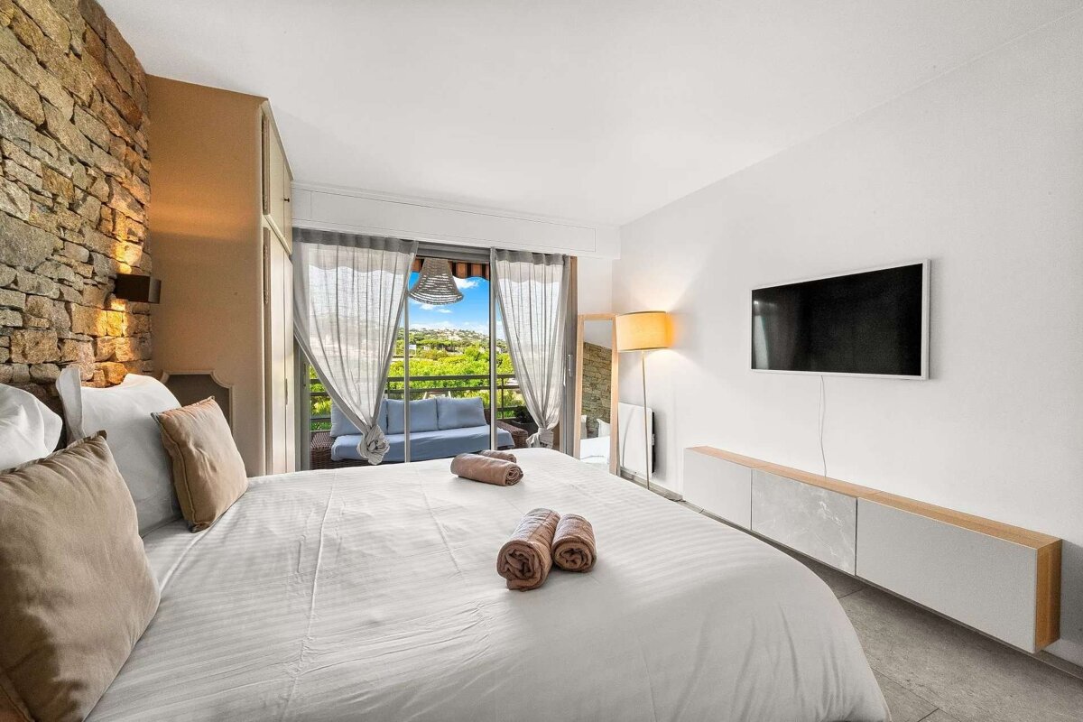 A stylish bedroom features a large bed, stone accent wall, TV, and a balcony overlooking lush green scenery.