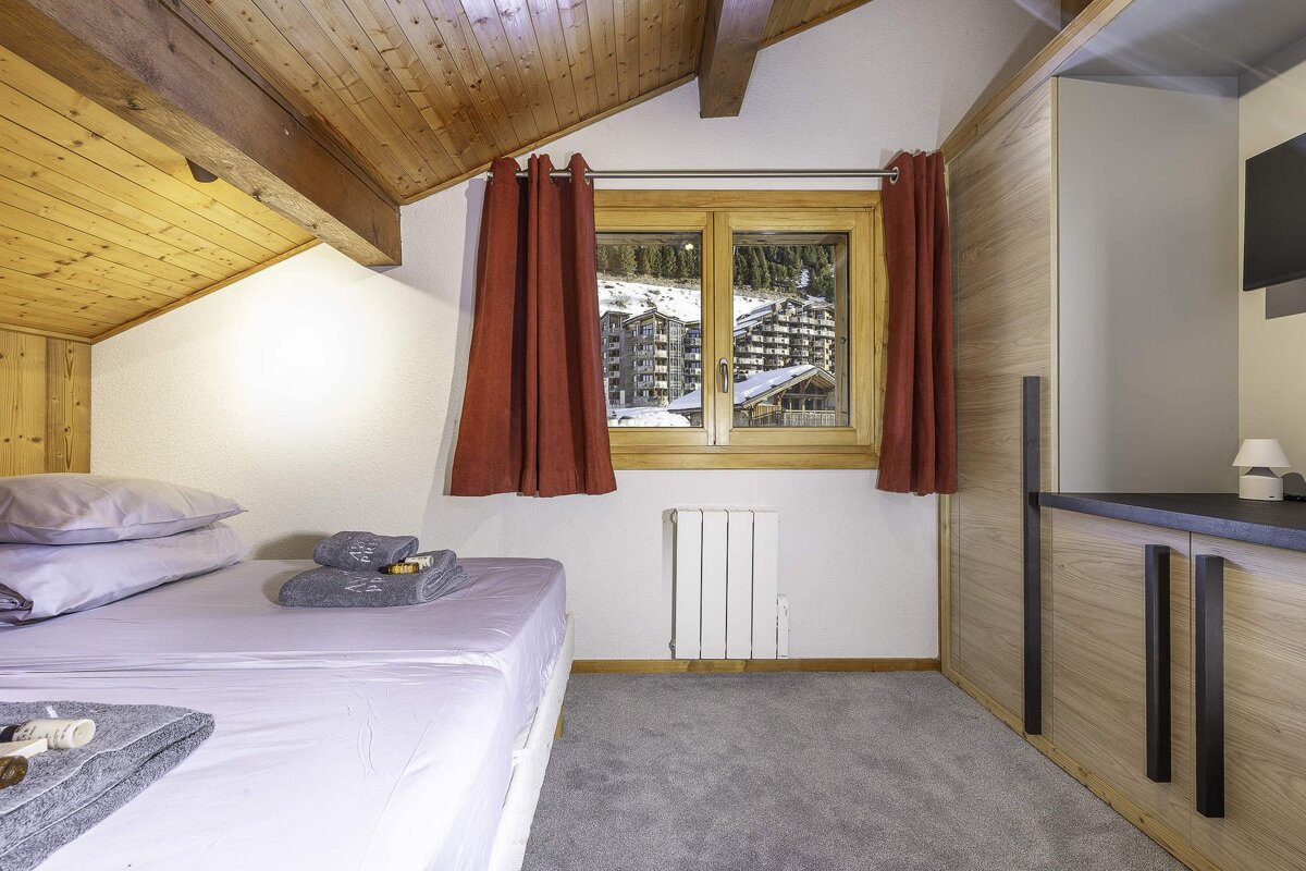 Cozy bedroom with a bed, red curtains, wooden ceiling, and a window overlooking a snowy mountain village with ski chalets.