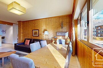Wood-paneled chalet room features a dining table, sofa, and bright windows with mountain & village views. Warm, inviting atmosphere.