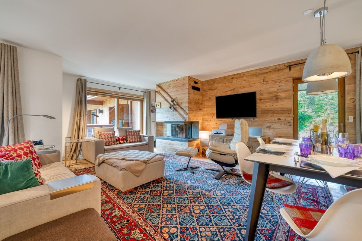 Stylish chalet living/dining room features wood walls, a modern fireplace, vibrant patterned rug, comfy sofas, and a set dining table.
