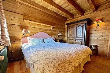A cozy rustic bedroom with all-wooden walls and ceiling, featuring a large bed with a pink headboard and an antique wooden wardrobe.