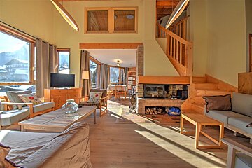 Cozy chalet living room with a fireplace, wooden stairs, and large windows revealing a snowy mountain view. Bright and inviting.