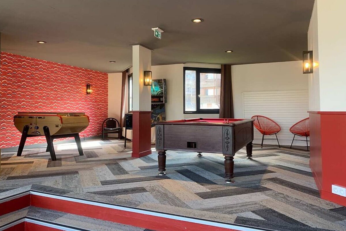 A vibrant game room with a foosball table, pool table, and pinball machine. Features red wavy walls, red accents, and a geometric floor.
