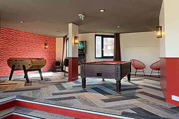 A vibrant game room with a foosball table, pool table, and pinball machine. Features red wavy walls, red accents, and a geometric floor.