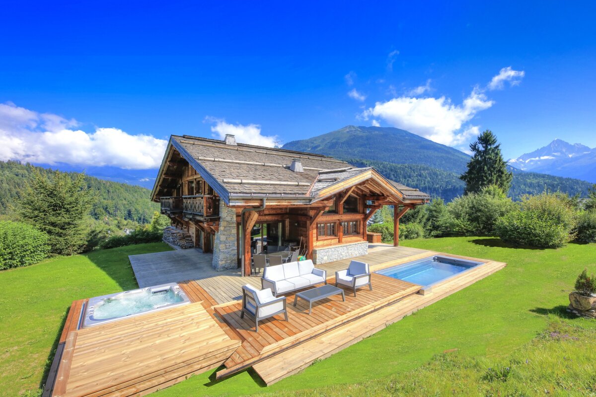 Stunning wooden chalet with a pool, hot tub, and large deck, nestled in a vibrant green mountain landscape under a clear blue sky.