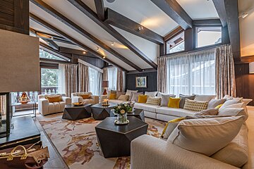 A luxurious living room features high beamed ceilings, large windows with sheer curtains, plush white sofas with yellow accents, and geometric coffee tables on a floral rug.